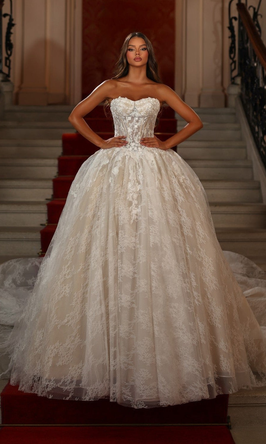 A woman stands on a red-carpeted staircase wearing the Alyce 7163 long white dress in Ivory/Sand, featuring a corset bodice and full skirt, with her hands on her hips.